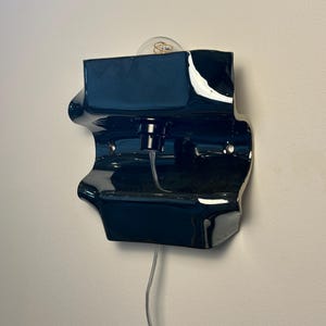 May include: A glossy dark navy blue ceramic wall sconce with a sculptural, undulating form. The fixture features a central light socket holding a clear glass bulb, with a transparent electrical cord extending downward from the base of the unit.