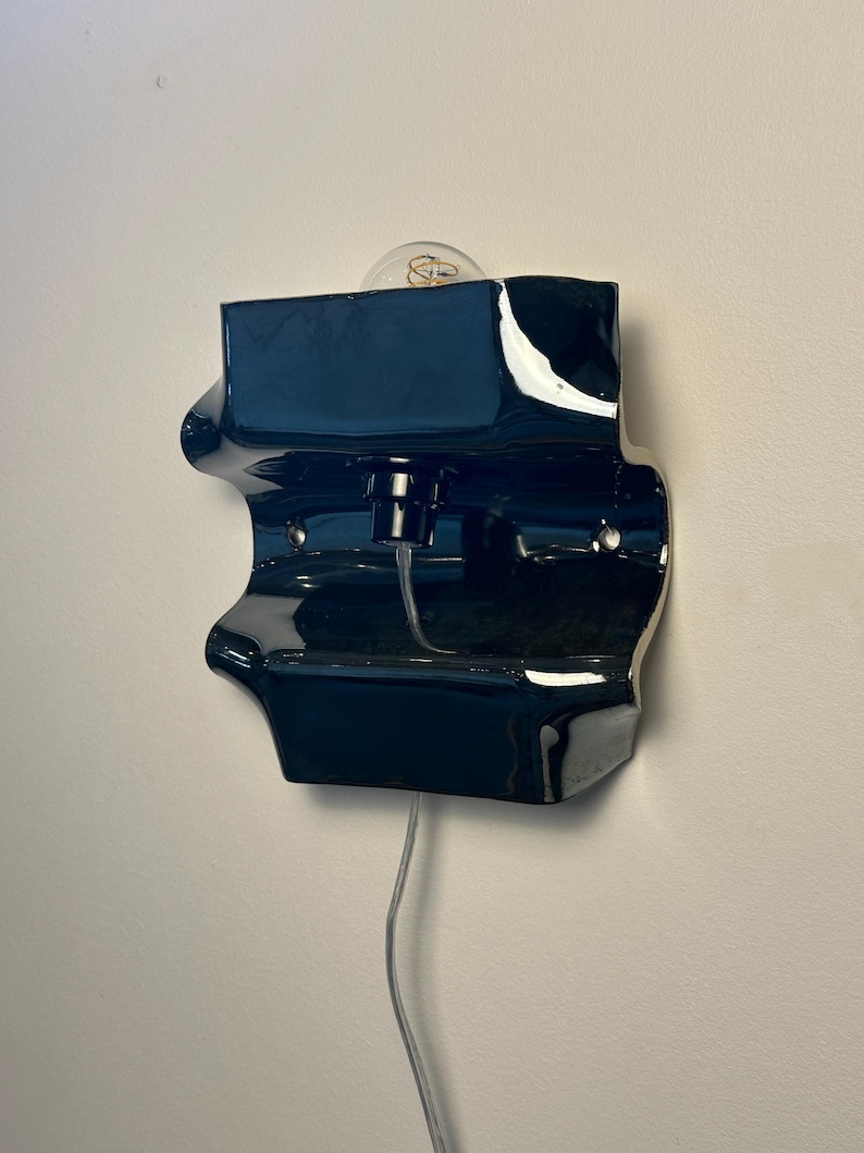May include: A glossy dark navy blue ceramic wall sconce with a sculptural, undulating form. The fixture features a central light socket holding a clear glass bulb, with a transparent electrical cord extending downward from the base of the unit.