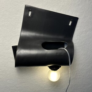 Puede incluir: Un aplique de pared negro mate con un dise&ntilde;o de l&aacute;mina de metal curvada y plegada con dos ranuras de montaje rectangulares en la parte superior. Un recorte horizontal central sostiene una bombilla esf&eacute;rica esmerilada con cable transparente.