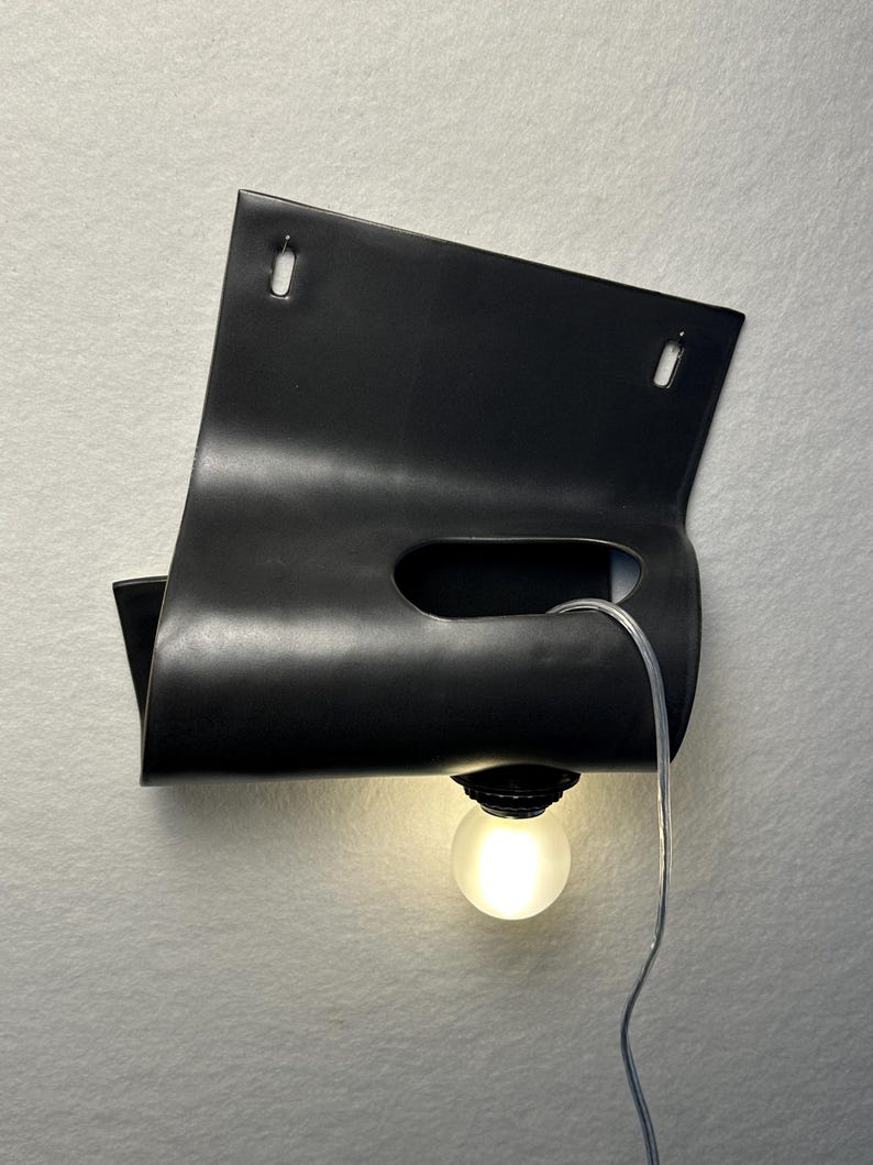 Puede incluir: Un aplique de pared negro mate con un dise&ntilde;o de l&aacute;mina de metal curvada y plegada con dos ranuras de montaje rectangulares en la parte superior. Un recorte horizontal central sostiene una bombilla esf&eacute;rica esmerilada con cable transparente.