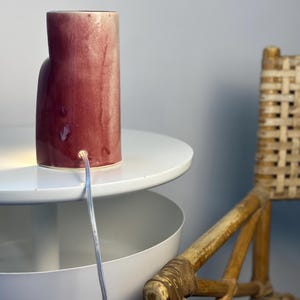 May include: A cylindrical ceramic table lamp with a glossy, mottled deep red and cream glaze. The lamp features a clear electrical cord extending from a circular port near the base, emitting a warm light from the bottom edge of the structure.