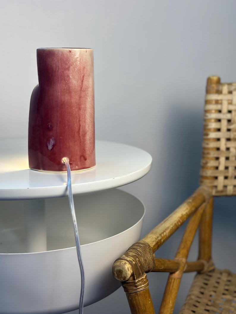 May include: A cylindrical ceramic table lamp with a glossy, mottled deep red and cream glaze. The lamp features a clear electrical cord extending from a circular port near the base, emitting a warm light from the bottom edge of the structure.
