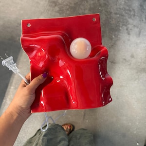 May include: A glossy red ceramic wall lamp with a wavy, undulating surface and a single round white light bulb integrated into the form. The fixture features two circular mounting holes at the top edge and includes a clear electrical power cord.