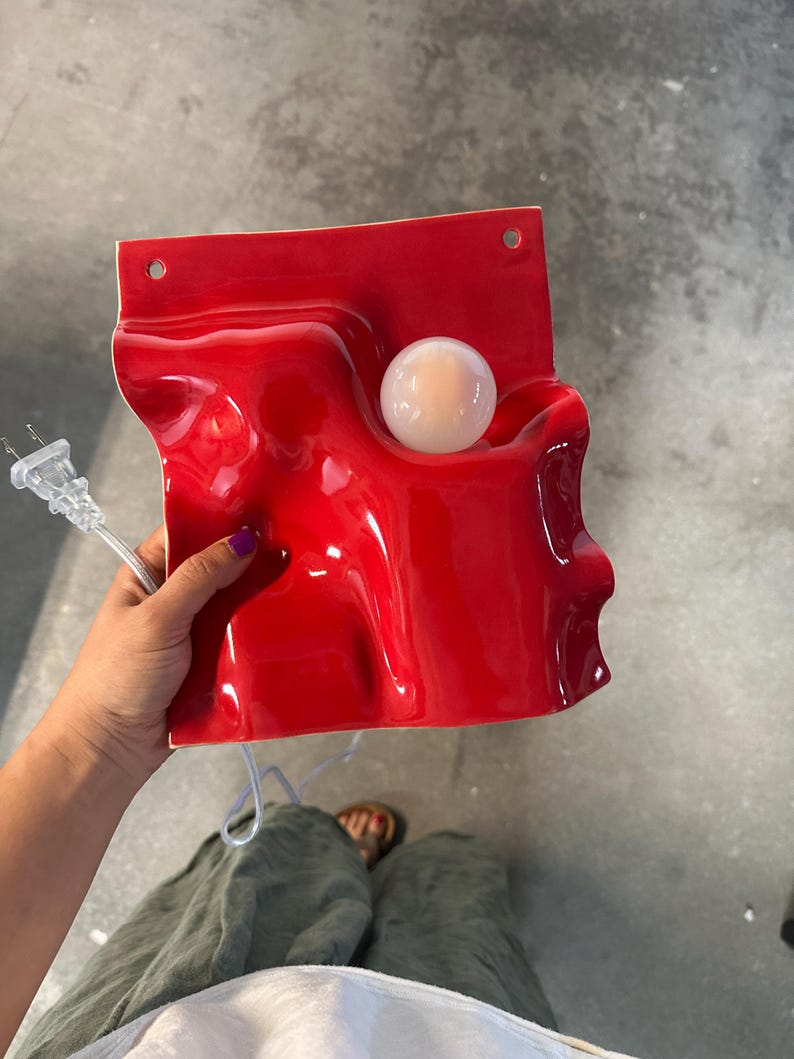 May include: A glossy red ceramic wall lamp with a wavy, undulating surface and a single round white light bulb integrated into the form. The fixture features two circular mounting holes at the top edge and includes a clear electrical power cord.