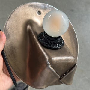 May include: A wall-mounted lamp featuring a circular, metallic bronze-toned backplate with a folded, sculptural design. A black socket is centered on the plate, holding a small, round, frosted white light bulb. A black power cord extends from the side.