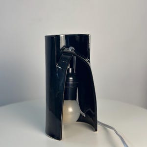 May include: A black ceramic table lamp with a curved, cylindrical structure. The lamp features a central opening that houses a black socket and a round, frosted white light bulb. A light-colored electrical cord extends from the base.