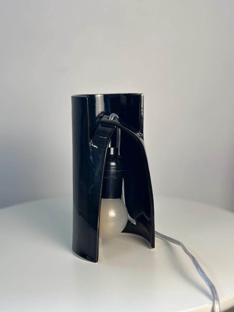 May include: A black ceramic table lamp with a curved, cylindrical structure. The lamp features a central opening that houses a black socket and a round, frosted white light bulb. A light-colored electrical cord extends from the base.