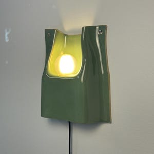 May include: A sage green ceramic wall sconce with a glossy finish and a curved, organic shape. The fixture features a central cutout housing a glowing, spherical light bulb. A black power cord extends from the bottom, and two small mounting holes are visible at the top corners of the structure.