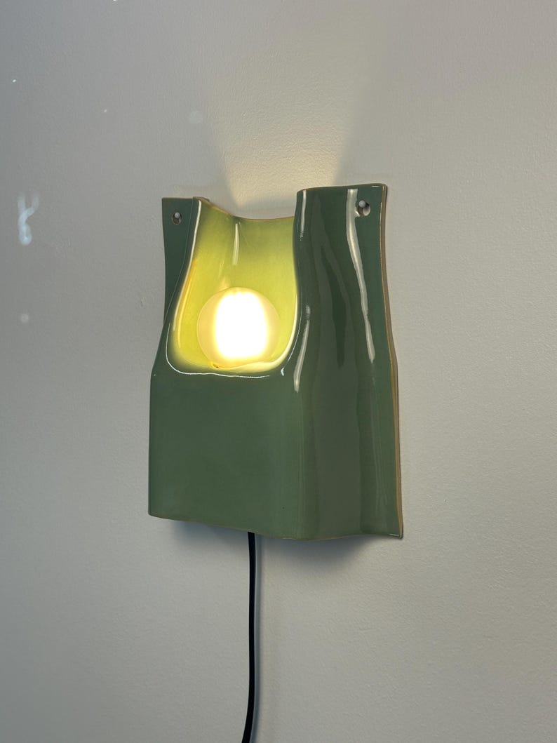 May include: A sage green ceramic wall sconce with a glossy finish and a curved, organic shape. The fixture features a central cutout housing a glowing, spherical light bulb. A black power cord extends from the bottom, and two small mounting holes are visible at the top corners of the structure.