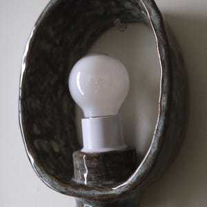 May include: A ceramic wall sconce with a bulb. The sconce has a dark brown, oval-shaped frame with a textured surface. A white light bulb is mounted in the center, and a clear cord hangs below.