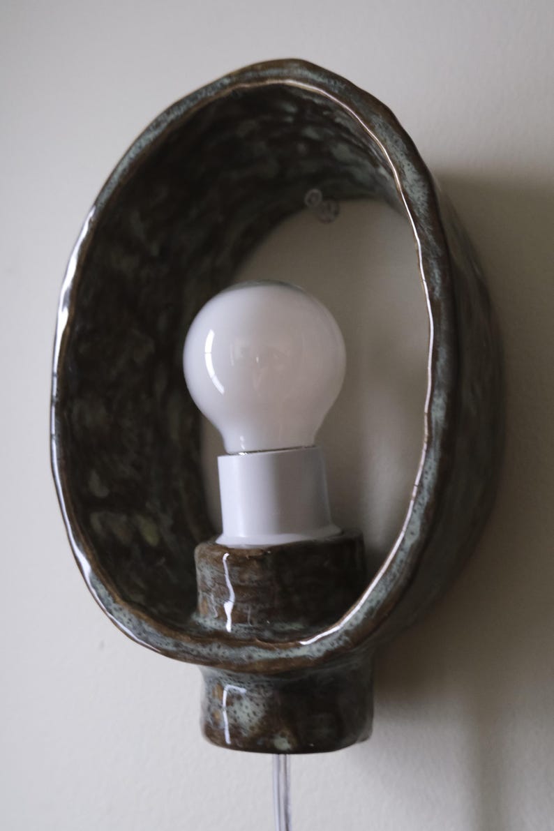May include: A ceramic wall sconce with a bulb. The sconce has a dark brown, oval-shaped frame with a textured surface. A white light bulb is mounted in the center, and a clear cord hangs below.