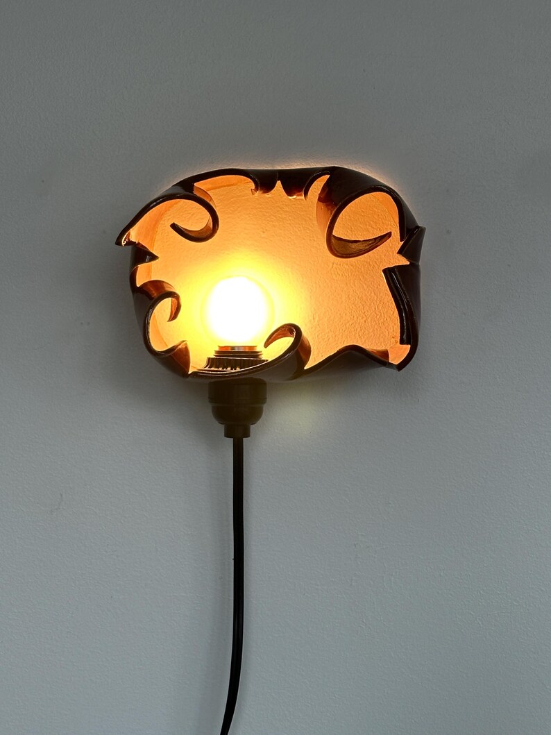 May include: A decorative wall sconce with a warm, glowing light. The sconce has a dark brown, organic shape with a light bulb in the center. The light is surrounded by a cutout design. A black cord extends downward.