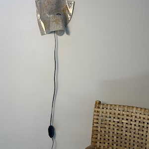 May include: A wall-mounted ceramic sconce with a speckled beige and grey finish, featuring an irregular, folded shape. The light fixture includes a single exposed light bulb and a long black power cord with an inline switch hanging vertically.