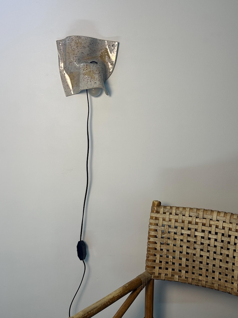 May include: A wall-mounted ceramic sconce with a speckled beige and grey finish, featuring an irregular, folded shape. The light fixture includes a single exposed light bulb and a long black power cord with an inline switch hanging vertically.
