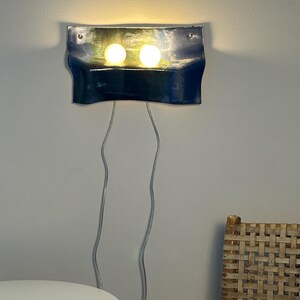 May include: A wall-mounted lamp featuring a curved, glossy dark blue rectangular panel with two illuminated round white bulbs. Two thin, light-colored electrical cords hang vertically from the bottom of the fixture, which is secured by two silver screws.