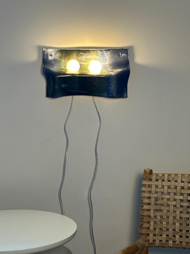 May include: A wall-mounted lamp featuring a curved, glossy dark blue rectangular panel with two illuminated round white bulbs. Two thin, light-colored electrical cords hang vertically from the bottom of the fixture, which is secured by two silver screws.