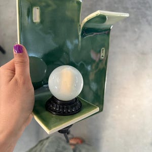 May include: A wall-mounted ceramic sconce featuring a glossy, deep green finish with a rectangular backplate and a small, protruding shelf. A round, frosted white glass light bulb is secured in a black, fluted base centered on the shelf.