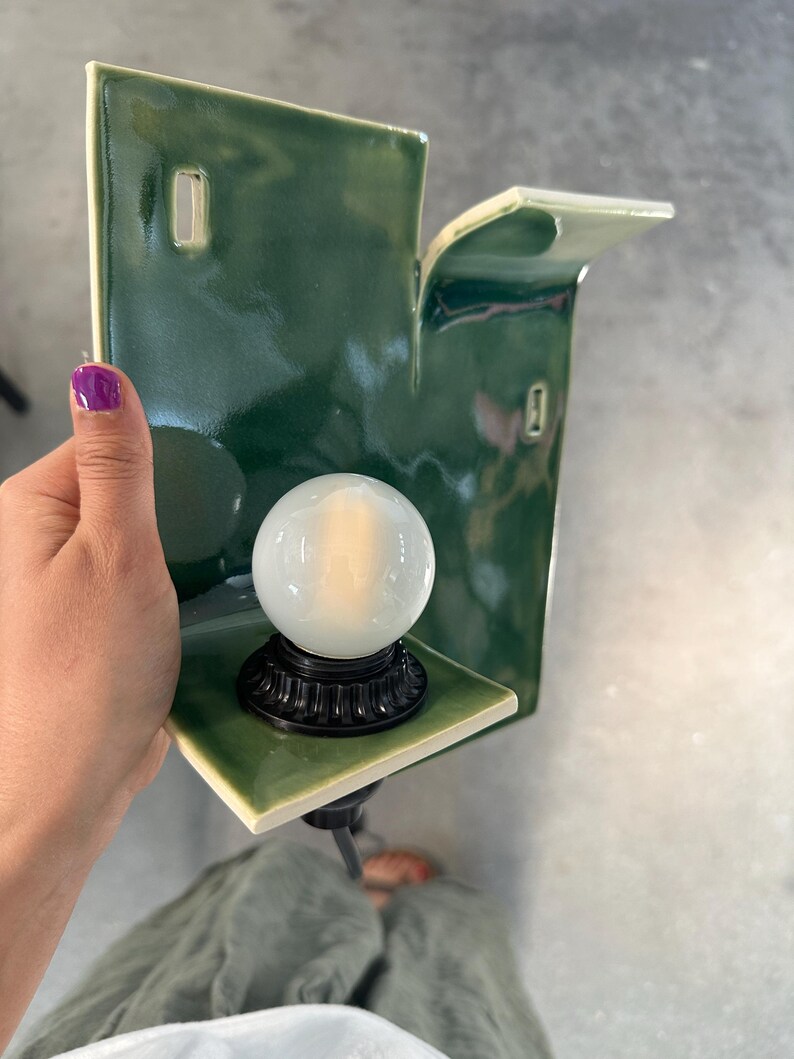 May include: A wall-mounted ceramic sconce featuring a glossy, deep green finish with a rectangular backplate and a small, protruding shelf. A round, frosted white glass light bulb is secured in a black, fluted base centered on the shelf.