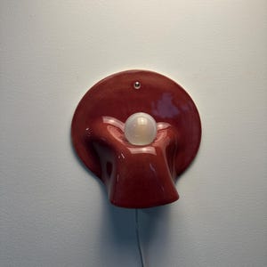 May include: A wall-mounted lamp featuring a glossy, deep red ceramic base with a fluid, organic, shell-like shape. A single, spherical, frosted white light bulb is centred within the curved, recessed middle section of the ceramic fixture.