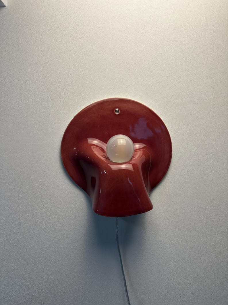 May include: A wall-mounted lamp featuring a glossy, deep red ceramic base with a fluid, organic, shell-like shape. A single, spherical, frosted white light bulb is centred within the curved, recessed middle section of the ceramic fixture.