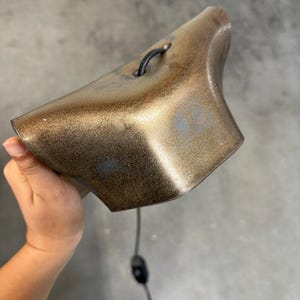 Pode incluir: Um sino de cor bronze met&aacute;lico com um acabamento texturizado e salpicado. O sino apresenta uma forma curva e alargada, incluindo um pequeno la&ccedil;o preto no topo para pendurar. Um fio el&eacute;trico preto fino com um interruptor sai da parte inferior.