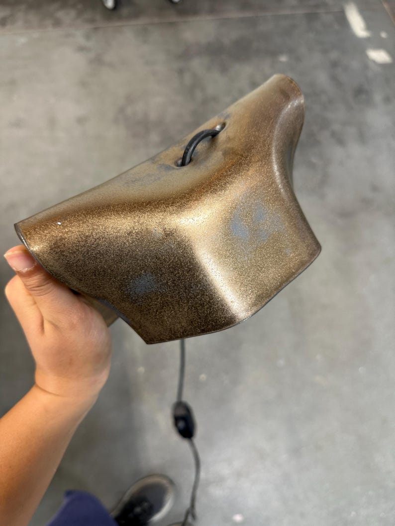 Pode incluir: Um sino de cor bronze met&aacute;lico com um acabamento texturizado e salpicado. O sino apresenta uma forma curva e alargada, incluindo um pequeno la&ccedil;o preto no topo para pendurar. Um fio el&eacute;trico preto fino com um interruptor sai da parte inferior.