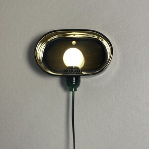 K&ouml;nnte beinhalten: Eine ovale Wandleuchte mit schwarzem Rahmen und goldfarbenem Innenraum. Sie beherbergt eine runde, wei&szlig;e Gl&uuml;hbirne. Ein gr&uuml;ner Sockel und ein schwarzes Kabel gehen von unten ab. Die Leuchte ist an einer grauen Wand befestigt.