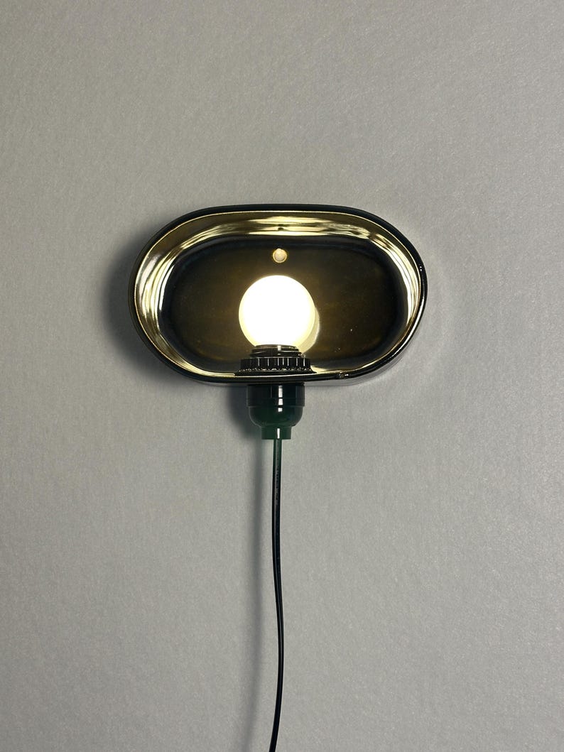 K&ouml;nnte beinhalten: Eine ovale Wandleuchte mit schwarzem Rahmen und goldfarbenem Innenraum. Sie beherbergt eine runde, wei&szlig;e Gl&uuml;hbirne. Ein gr&uuml;ner Sockel und ein schwarzes Kabel gehen von unten ab. Die Leuchte ist an einer grauen Wand befestigt.
