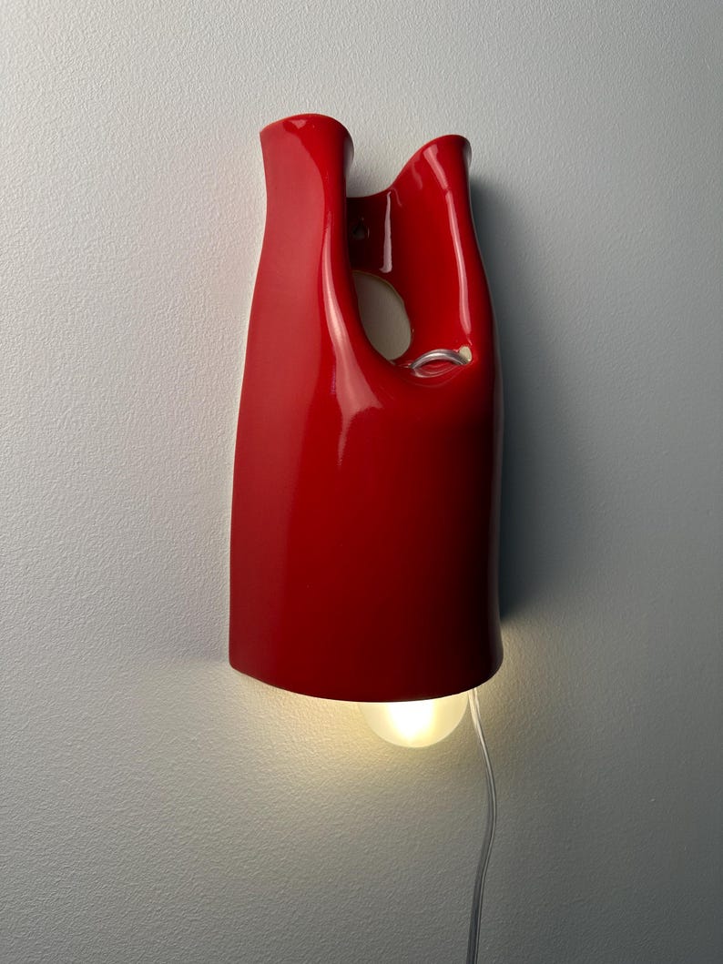May include: A glossy red wall-mounted lamp with a smooth, organic, and asymmetrical silhouette. The fixture features a central cutout and a downward-facing light bulb that emits a soft glow, connected by a thin, transparent electrical cord.