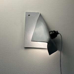 May include: A white ceramic wall lamp with a folded, sheet-like design. The fixture features a small circular hole at the top corner for mounting, a black socket attached to the side, and a thin gray power cord extending downward from the base.