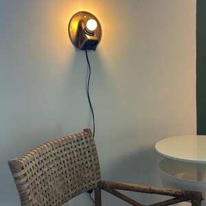 May include: A circular wall-mounted lamp with a metallic bronze finish and a single glowing white bulb. The fixture features a black power cord extending downward. The lamp emits a warm, soft light against the wall, illuminating the surrounding area.