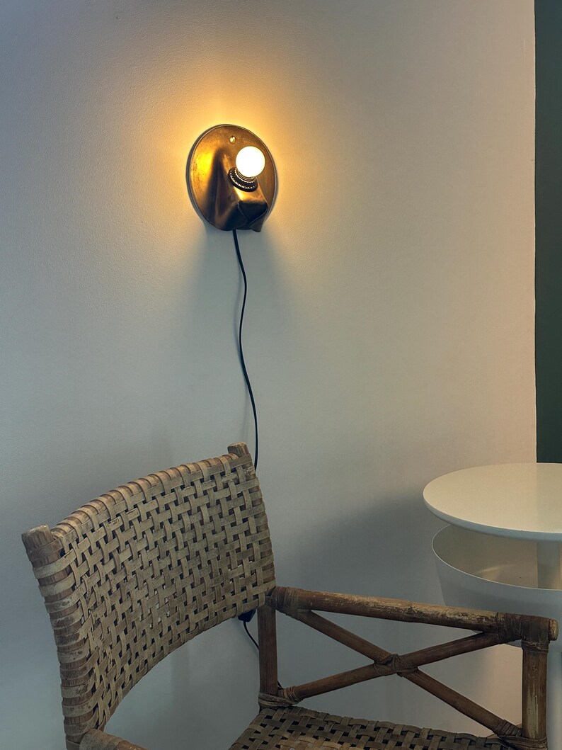 May include: A circular wall-mounted lamp with a metallic bronze finish and a single glowing white bulb. The fixture features a black power cord extending downward. The lamp emits a warm, soft light against the wall, illuminating the surrounding area.