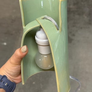 May include: A sage green ceramic lamp with a curved, open-sided cylindrical body. The structure features a central cutout holding a white light socket and a clear glass light bulb. A thin, transparent electrical cord extends from the top.