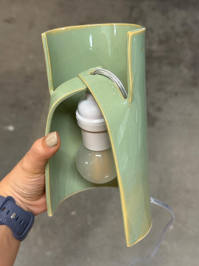 May include: A sage green ceramic lamp with a curved, open-sided cylindrical body. The structure features a central cutout holding a white light socket and a clear glass light bulb. A thin, transparent electrical cord extends from the top.