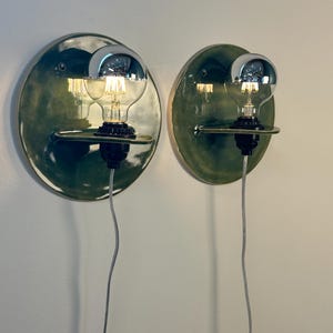 May include: Two wall-mounted light fixtures featuring circular, glossy dark green ceramic backplates. Each fixture has a small horizontal shelf holding a black socket with a clear glass light bulb that has a reflective silver-toned top cap.