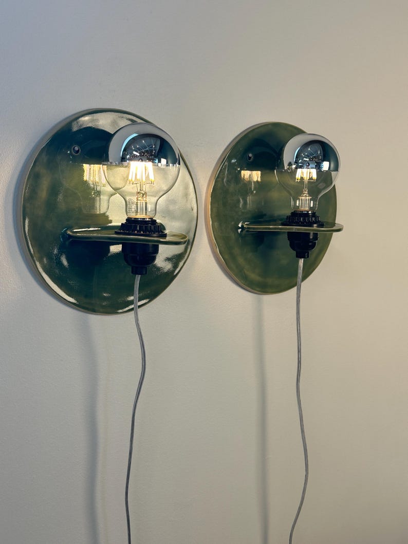 May include: Two wall-mounted light fixtures featuring circular, glossy dark green ceramic backplates. Each fixture has a small horizontal shelf holding a black socket with a clear glass light bulb that has a reflective silver-toned top cap.