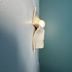 May include: A wall-mounted light fixture featuring a curved, off-white ceramic shade with a black speckled pattern. The fixture has a wavy, organic shape and holds a single glowing, spherical light bulb that emits a warm, soft light.
