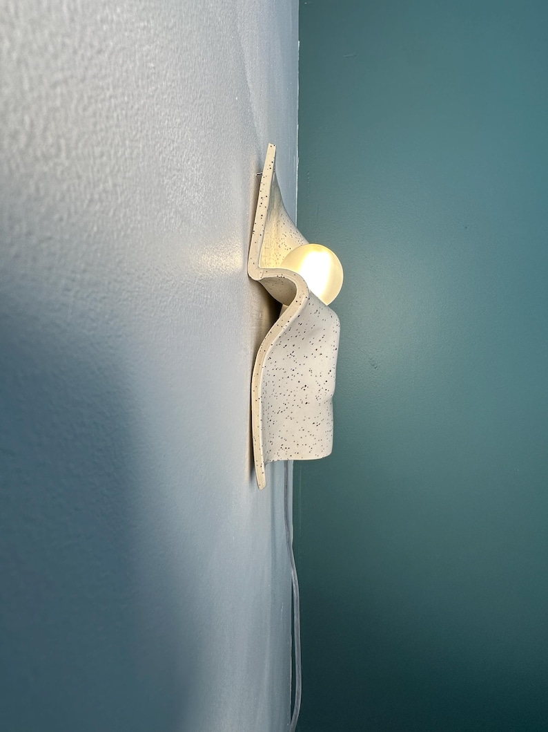 May include: A wall-mounted light fixture featuring a curved, off-white ceramic shade with a black speckled pattern. The fixture has a wavy, organic shape and holds a single glowing, spherical light bulb that emits a warm, soft light.