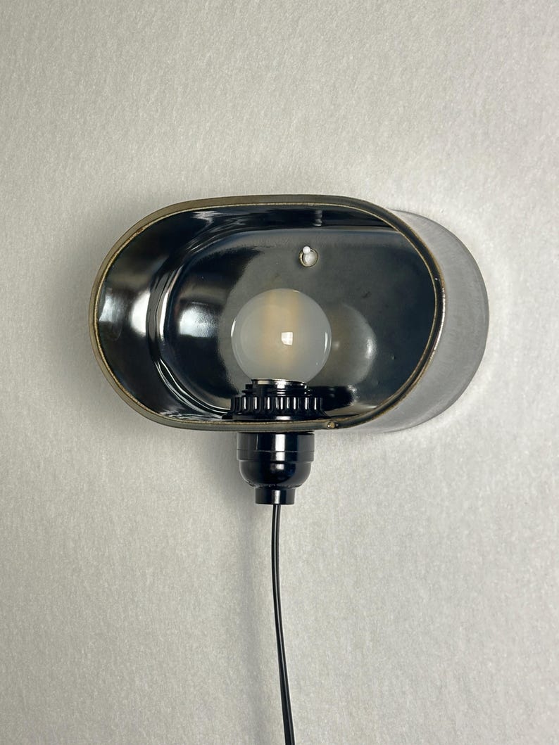 K&ouml;nnte beinhalten: Eine Wandleuchte mit einem verchromten, ovalen Schirm. Die Leuchte beherbergt eine mattierte Gl&uuml;hbirne und einen schwarzen Sockel mit Netzkabel. Das Design ist minimalistisch und industriell, passend f&uuml;r verschiedene Einrichtungsstile.