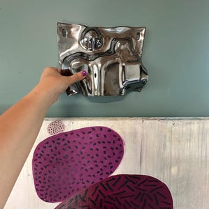 Pu&ograve; includere: Una placca da parete in metallo argentato, ondulata e riflettente, con una superficie liscia simile a un liquido e due piccoli fori di montaggio negli angoli superiori, posizionata sopra una tela con due grandi forme ovali viola con motivi a trattini.