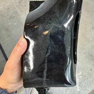May include: A glossy black ceramic vessel with a rectangular, slightly curved silhouette and a smooth, reflective finish. The object features a dark, mottled glaze with subtle hints of grey and a small, circular black spout at the base.