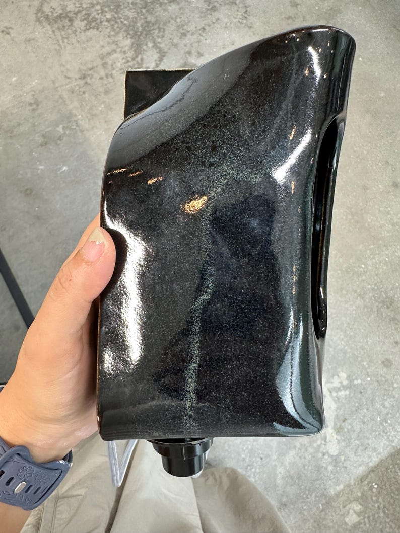 May include: A glossy black ceramic vessel with a rectangular, slightly curved silhouette and a smooth, reflective finish. The object features a dark, mottled glaze with subtle hints of grey and a small, circular black spout at the base.