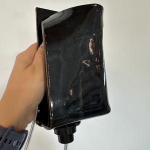 May include: A glossy black ceramic wall sconce with a rectangular, slightly curved silhouette. The fixture features a dark, reflective glaze with subtle metallic undertones and a black circular socket at the base for a light bulb and cord.