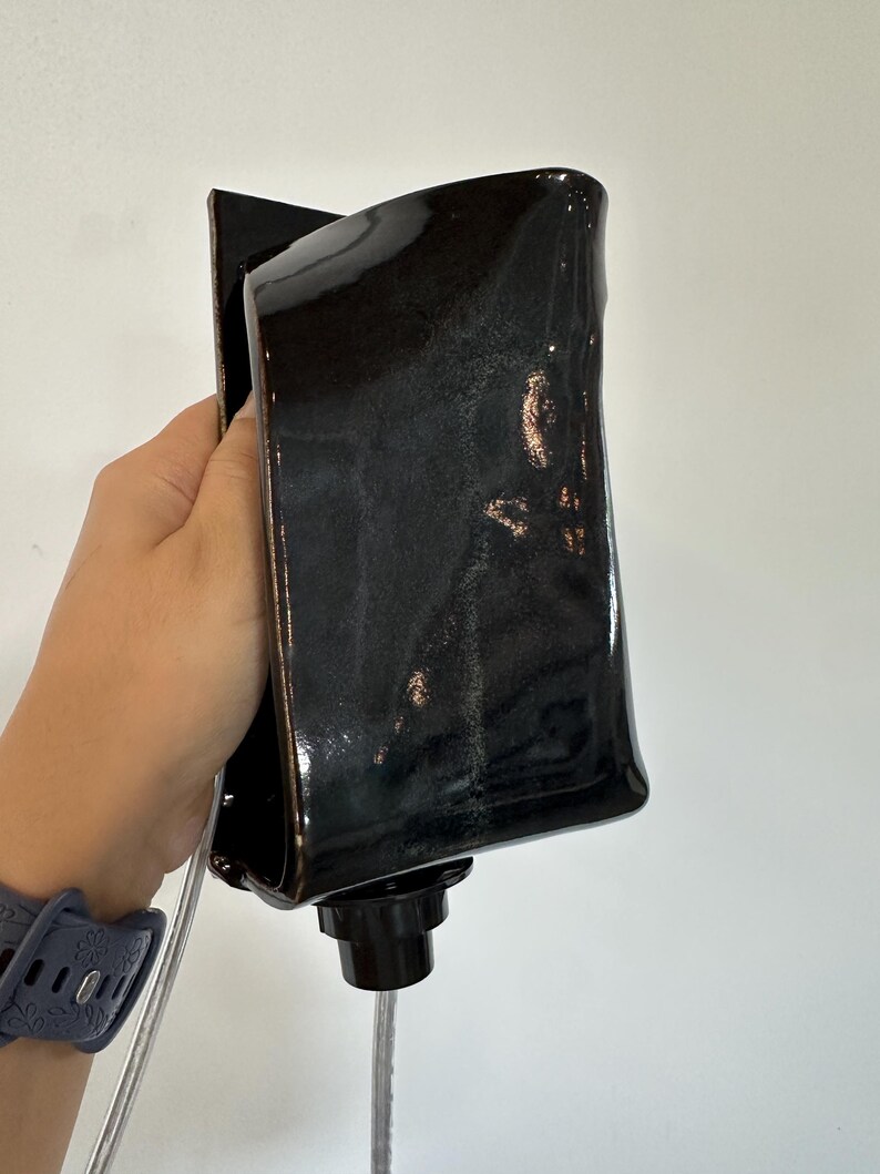 May include: A glossy black ceramic wall sconce with a rectangular, slightly curved silhouette. The fixture features a dark, reflective glaze with subtle metallic undertones and a black circular socket at the base for a light bulb and cord.