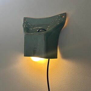 May include: A wall-mounted ceramic light fixture in a speckled sage green glaze. The sconce features a flared, rectangular shape with two mounting holes at the top corners and a black power cord extending from the bottom, emitting a warm glow.