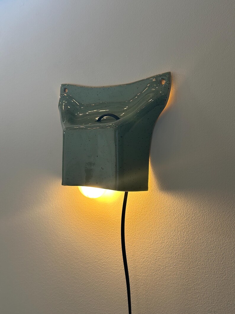 May include: A wall-mounted ceramic light fixture in a speckled sage green glaze. The sconce features a flared, rectangular shape with two mounting holes at the top corners and a black power cord extending from the bottom, emitting a warm glow.