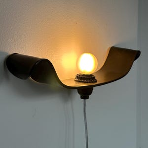 May include: A wall-mounted, bronze-colored sconce with a curved, ribbon-like design. The sconce holds a round, illuminated light bulb. The fixture is attached to the wall with a cord.