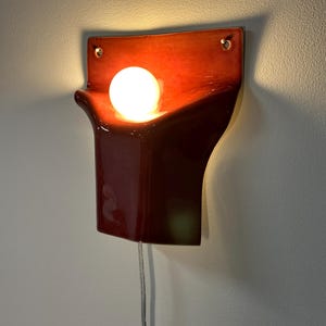 Pode incluir: Um candeeiro de parede em cer&acirc;mica com um acabamento vidrado vermelho profundo e brilhante. A pe&ccedil;a apresenta uma forma escultural curva com dois orif&iacute;cios de montagem nos cantos superiores. Uma l&acirc;mpada branca redonda brilha no centro.