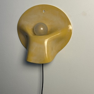 May include: A wall-mounted lamp featuring a smooth, yellow, organic-shaped ceramic shade with a glossy finish. A single, round, frosted white light bulb is centered within the concave form. A thin black power cord extends from the bottom of the fixture.