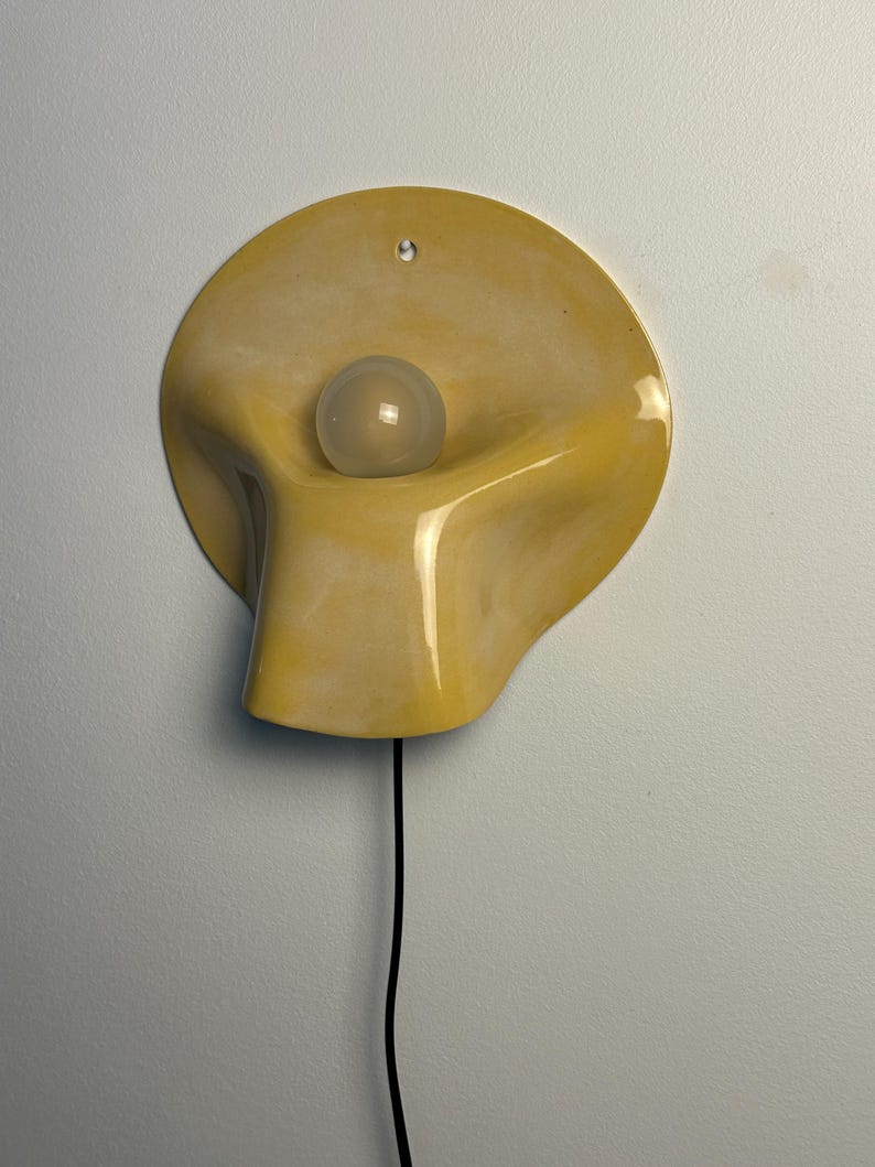 May include: A wall-mounted lamp featuring a smooth, yellow, organic-shaped ceramic shade with a glossy finish. A single, round, frosted white light bulb is centered within the concave form. A thin black power cord extends from the bottom of the fixture.